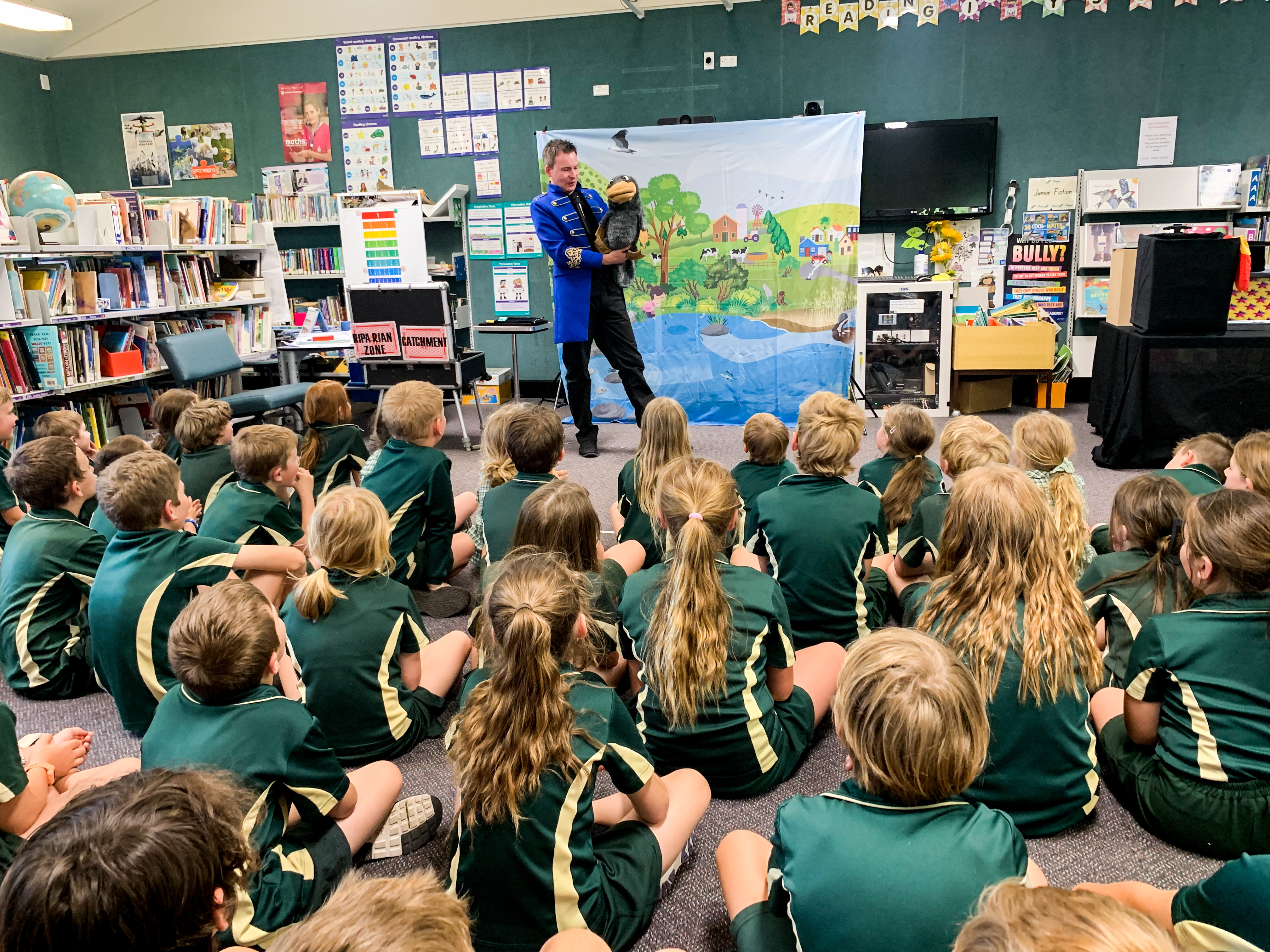 A classroom of children sitting on the floor listening to a man in a blue coat with a platypus puppet